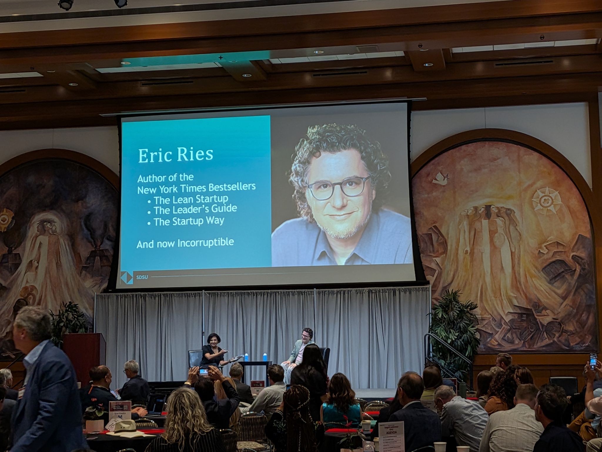 Wide shot of Eric Ries on stage with interviewer Vidya Dinamani at SDSU, audience in foreground, large Eric Ries title slide projected behind them